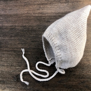 May include: A light gray knitted baby bonnet with a pointed top and attached white ties. The bonnet is made of a soft, textured yarn and is shown on a dark wood surface. A cozy accessory for newborns.
