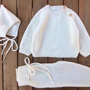 Könnte beinhalten: Ein weißes Strick-Baby-Outfit-Set bestehend aus einem Pullover, einer Hose und einer Mütze. Der Pullover hat einen runden Ausschnitt und Knopfverschlüsse an der Schulter. Die Hose hat einen Kordelzugbund. Die Mütze hat eine spitze Oberseite und Bänder unter dem Kinn.