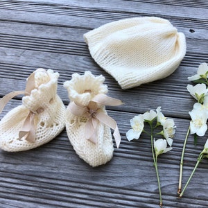 Puede incluir: Un conjunto de botines de beb&eacute; de punto blanco con lazos de sat&eacute;n beige y un gorro de punto blanco.