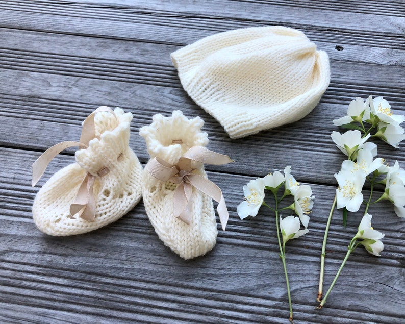 Puede incluir: Un conjunto de botines de beb&eacute; de punto blanco con lazos de sat&eacute;n beige y un gorro de punto blanco.