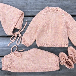 May include: A knitted baby outfit in a pale pink colour. The set includes a sweater, trousers, a hat, and booties.