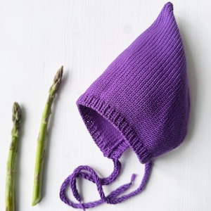 Peut inclure: Un bonnet de bébé violet tricoté avec un haut pointu et des liens attachés. Deux asperges vertes sont positionnées à gauche du bonnet sur une surface blanche. Le bonnet est fait d'un fil doux et texturé.