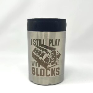 May include: A stainless steel can cooler with a black rubber top. The cooler has the text "I STILL PLAY WITH BLOCKS" and a graphic of an engine block.