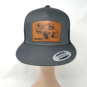 May include: A gray snapback hat with a black mesh back. The front features a brown leather patch with a motorcycle graphic and the word "Spyder". A white and black sticker is on the brim.