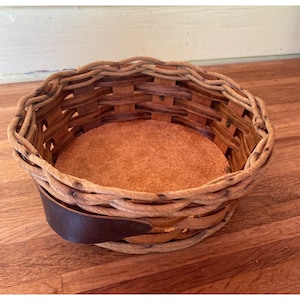 May include: A round woven basket featuring light brown wooden splints and a thick, dark brown leather strap attached to the side. The basket has a flat, circular base made of a textured, light brown material and a braided rim along the top edge.