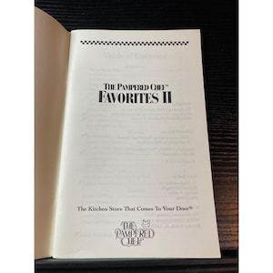 May include: Open book with the title "THE PAMPERED CHEF FAVORITES II" in bold black letters. The text "The Kitchen Store That Comes To Your Door" is below the title. The Pampered Chef logo is at the bottom of the page.