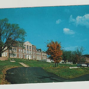 May include: Vintage postcard featuring a brick building with white columns and a sloped roof, set against a bright blue sky with fluffy clouds. A road curves in front of the building, with trees and grass.