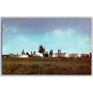 May include: Vintage postcard featuring an industrial landscape with oil storage tanks and smokestacks against a blue sky. The foreground shows a field of tall grass. The image has a retro aesthetic.