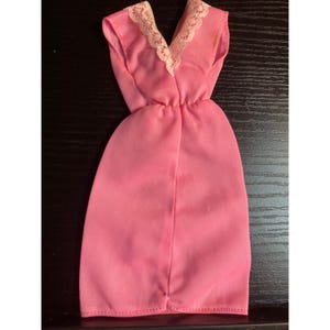 May include: A vintage pink doll dress with a V-neckline trimmed with cream-colored lace. The sleeveless dress has a gathered waist and a simple, straight skirt. The dress is made of a lightweight fabric.