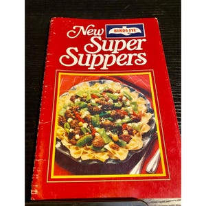 May include: A vintage cookbook titled "New Super Suppers" by Birds Eye. The cover is red with a yellow border and features a photo of a pasta dish with meat, green beans, and corn. The cookbook is open on a dark surface.