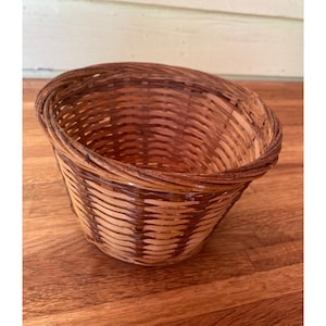 May include: A round, open-top woven basket constructed from thin, flexible strips of wood. The basket features a natural light brown color with darker brown vertical accents woven throughout the sides, ending in a thick, reinforced circular rim.