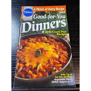 May include: A Pillsbury Classic Cookbooks book titled "Good-for-You Dinners" with a photo of a skillet meal. The cover includes text about Crock-Pot recipes and a vegetable fiesta skillet supper. The book is on a dark surface.