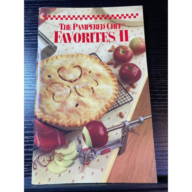 May include: A cookbook titled "The Pampered Chef Favorites II" with a pie on the cover. The pie has a decorative crust and is surrounded by apples, a cooling rack, and an apple peeler. The cover is cream-colored with red accents.