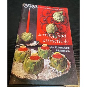 May include: A vintage cookbook titled "serving food attractively" by Florence Brobeck. The cover features artichokes, a red candle, and a serving dish with artichokes filled with red caviar and lemon slices. The background is red and dark brown.