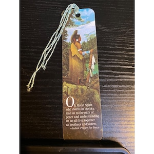 May include: Bookmark featuring an illustration of a Native American figure in traditional attire, set against a backdrop of a forest and sky. The bookmark includes a quote from the Indian Prayer for Peace and a light blue tassel.