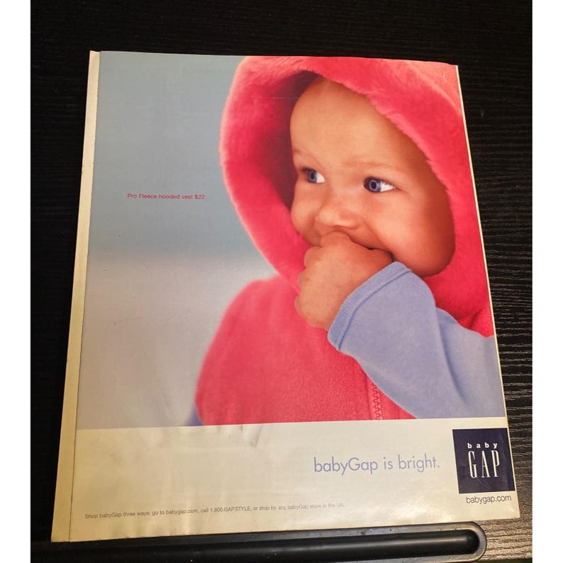 May include: A baby wearing a bright pink hooded fleece vest with a blue long-sleeved shirt underneath. The baby is looking to the side and has their hand in their mouth. The babyGap logo and the words "babyGap is bright" are at the bottom.