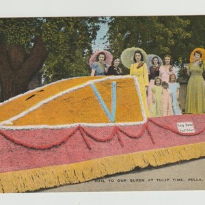 May include: Vintage postcard featuring a parade float decorated with yellow and pink flowers. The float has a large V-shaped design and is surrounded by women in dresses and large hats. Text reads "Hail to our Queen at Tulip Time, Pella, Iowa - 2."