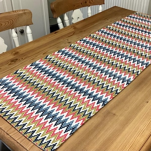 May include: A patterned table runner featuring a zig-zag design in red, blue, gold, and white. The runner is displayed on a wooden table, enhancing the dining space. The length of the runner is not visible, but it is a decorative item.