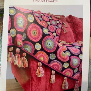 May include: A crochet blanket with a colourful circle pattern. The blanket is draped over a red sofa with tassels hanging from the edges. The blanket is titled "Magic Circles Crochet Blanket" and the designer is "Janie Crow".
