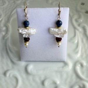 May include: Gold-coloured earrings with lever back fastenings. Each earring has a dark blue bead, a white, irregular pearl, clear crystal beads, and a cluster of red beads. A small white pearl hangs at the base.