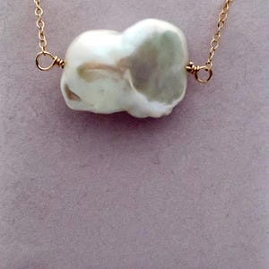 May include: A gold chain necklace featuring a large, irregularly shaped white pearl pendant. The pearl is attached to the chain with gold wire. The background is a soft, muted purple.