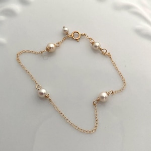 Puede incluir: Delicada pulsera de cadena dorada con pequeñas perlas redondas. La pulsera tiene un diseño sencillo con perlas espaciadas uniformemente a lo largo de la cadena. El cierre es un pequeño cierre de resorte redondo. La pulsera se muestra sobre una superficie blanca.