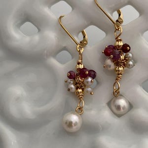 May include: A pair of gold earrings featuring clusters of small red and white beads, and a large white pearl. The earrings have a lever back closure.