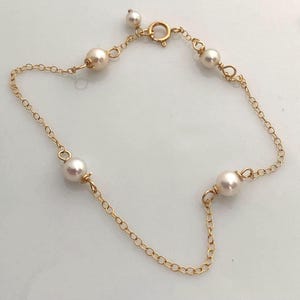 Pode incluir: Uma delicada pulseira dourada com um design de corrente e várias pérolas brancas. A pulseira tem um fecho e foi projetada para ser usada em volta do pulso. As pérolas são espaçadas uniformemente ao longo da corrente.