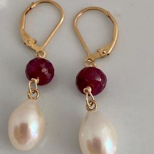 May include: A pair of gold-colored leverback earrings. Each earring features a faceted, deep red gemstone and a pear-shaped, creamy white pearl. The earrings are displayed against a light background.