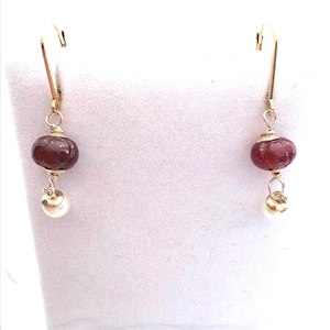May include: A pair of gold-colored earrings. Each earring features a faceted, deep red bead, a small white pearl, and a gold-colored hook. The earrings are displayed against a white background.