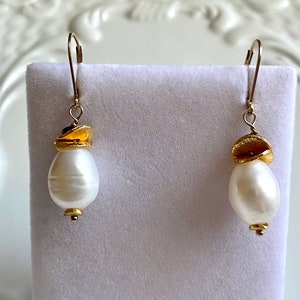 Puede incluir: Un par de pendientes de oro y perla blanca. Los pendientes presentan una sola perla blanca con un acento dorado.