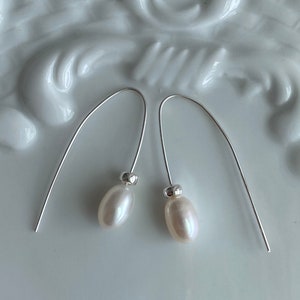 May include: A pair of elegant drop earrings featuring lustrous, oval-shaped pearls suspended from delicate, curved silver wires. The earrings rest on a white, textured surface, showcasing their simple yet sophisticated design.