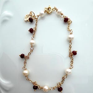 May include: A gold chain bracelet featuring alternating white pearls and small dark red faceted beads. The bracelet includes a circular spring ring clasp and a small gold leaf charm dangling near the closure, creating a delicate link design.