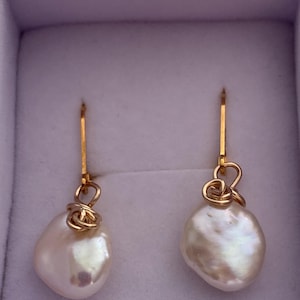 May include: A pair of gold-colored drop earrings featuring large, irregular-shaped white pearls. The pearls are suspended from gold wire hooks with a decorative gold wire wrap. The earrings are displayed in a light purple box.