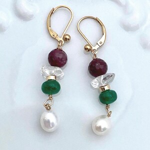 May include: A pair of gold-colored dangle earrings. Each earring features a deep red bead, a clear crystal, a green bead, and a white teardrop-shaped pearl. The earrings are displayed against a white background.