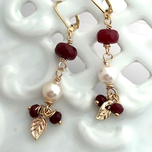 May include: A pair of gold-tone leverback earrings featuring a faceted deep red bead, a smooth white pearl, and a small gold-tone leaf charm at the base. The components are connected by gold-tone wire wrapping and small circular jump rings.