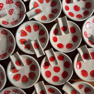 May include: A collection of ceramic plates and spoon rests with a strawberry theme. The plates are white with red strawberry designs, and the spoon rests are rectangular. Some plates feature mushroom designs.