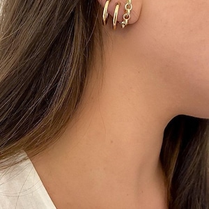 Puede incluir: Primer plano de una oreja adornada con tres pendientes de aro dorados y un pendiente de cadena. Los pendientes son de color dorado brillante y la persona tiene el pelo largo y castaño.