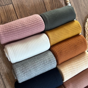 Peut inclure: Collection de tissus en tricot gaufré roulés, de différentes couleurs : rose, blanc, gris, bleu marine et jaune moutarde. Le tissu présente un motif texturé en forme de grille. Les rouleaux sont empilés, mettant en valeur la gamme de couleurs disponibles.