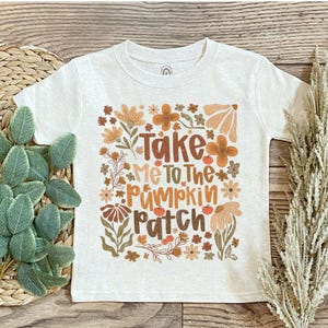 May include: A white t-shirt with the text "Take Me To The Pumpkin Patch" in brown and orange lettering. The shirt features floral and leaf designs in shades of brown, orange, and beige. The shirt is made of a soft material.
