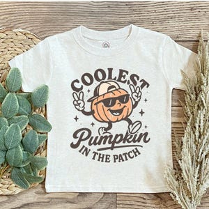 May include: A light gray t-shirt featuring a cartoon pumpkin wearing sunglasses and a backwards cap. The text on the shirt reads "COOLEST Pumpkin IN THE PATCH" in a brown, vintage-style font. The pumpkin is orange with a smiling face.