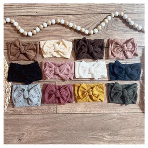 May include: A collection of twelve knotted headbands in various colors, including brown, cream, black, pink, white, navy, gray, burgundy, yellow, and green. The headbands are arranged on a wooden surface, with a beaded garland in the background.