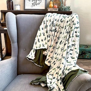 May include: A white blanket with a green cactus pattern draped over a gray armchair. The blanket has a green border. The chair is in front of a dark wooden cabinet with decorative items.