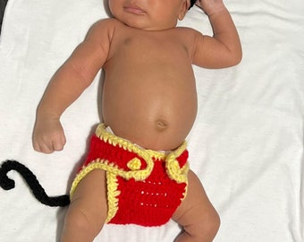 May include: A baby wearing a black crochet Mickey Mouse hat, red and yellow diaper cover, and white socks. The diaper cover has a black tail attached. The baby is lying on a white surface.