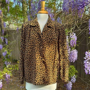May include: A brown and black leopard print button-up shirt with long sleeves. The shirt is on a mannequin in front of a background of purple wisteria flowers.