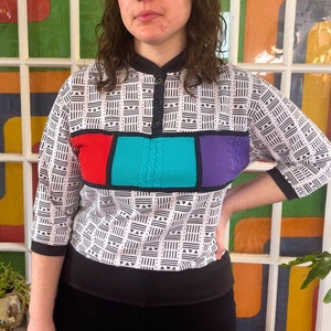 May include: A white and black geometric patterned shirt with a colorful geometric design across the chest. The design features red, turquoise, and purple.