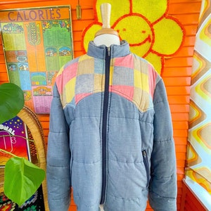 May include: A blue quilted jacket with a patchwork design on the shoulders and chest. The patchwork is made of pink, yellow, and blue squares. The jacket has a zipper closure and two pockets.
