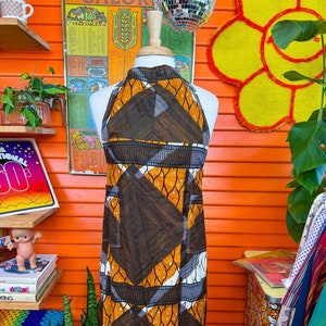 May include: A brown and orange halter dress with a geometric pattern. The dress has a high neckline and a fitted silhouette. The dress is on a mannequin against a bright orange wall.