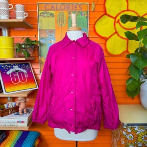 May include: A bright pink nylon jacket with a button-up front and long sleeves. The jacket is on a mannequin against a bright orange wall.