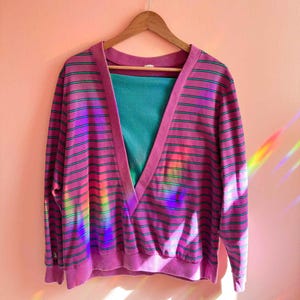 May include: A vintage, oversized, V-neck sweater in shades of pink and purple with horizontal stripes and teal accents. The sweater is hanging on a wooden hanger against a pink wall. A teal top is visible underneath.